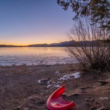 Una foto de una puesta de sol reflejada en el agua del lago Big Bear; en la orilla se ve un trozo de un trineo roto.