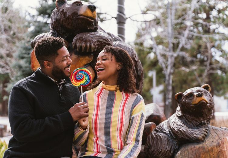 Una foto de una pareja compartiendo una piruleta con remolinos de colores del arcoíris, sentados frente a una estatua de un oso en Big Bear Lake Village.