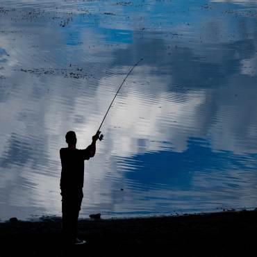 La silueta de un pescador se encuentra en la orilla del lago Big Bear y lanza un sedal al agua. El agua es azul