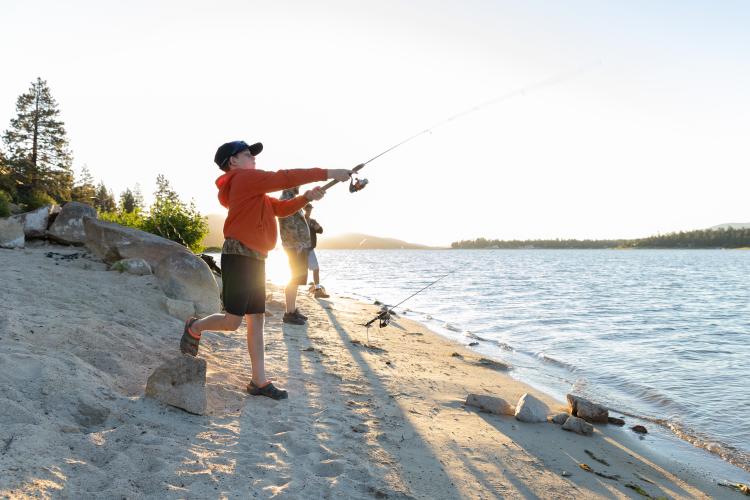 Un niño con chaqueta roja lanza un sedal al lago Big Bear mientras pesca en la playa con su familia.