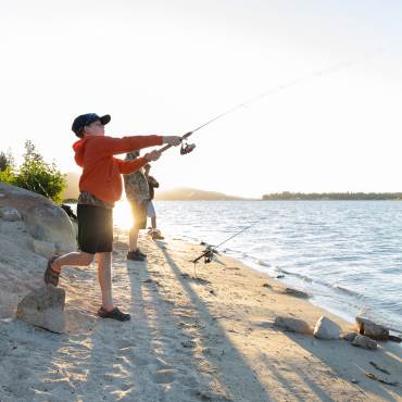 Un niño con chaqueta roja lanza un sedal al lago Big Bear mientras pesca en la playa con su familia.