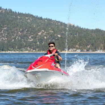 Una persona con chaleco salvavidas conduce una moto acuática roja por una masa de agua, creando olas en verano en Big Bear Lake, California.