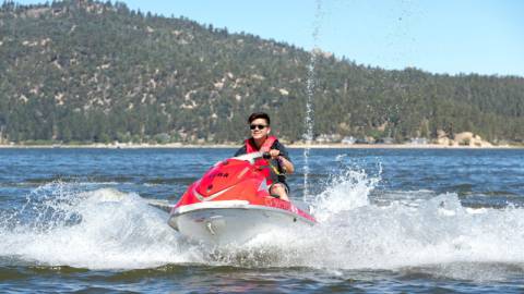 Una persona con chaleco salvavidas conduce una moto acuática roja por una masa de agua, creando olas en verano en Big Bear Lake, California.