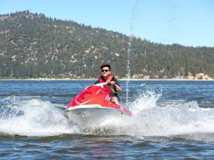 Una persona con chaleco salvavidas conduce una moto acuática roja por una masa de agua, creando olas en verano en Big Bear Lake, California.