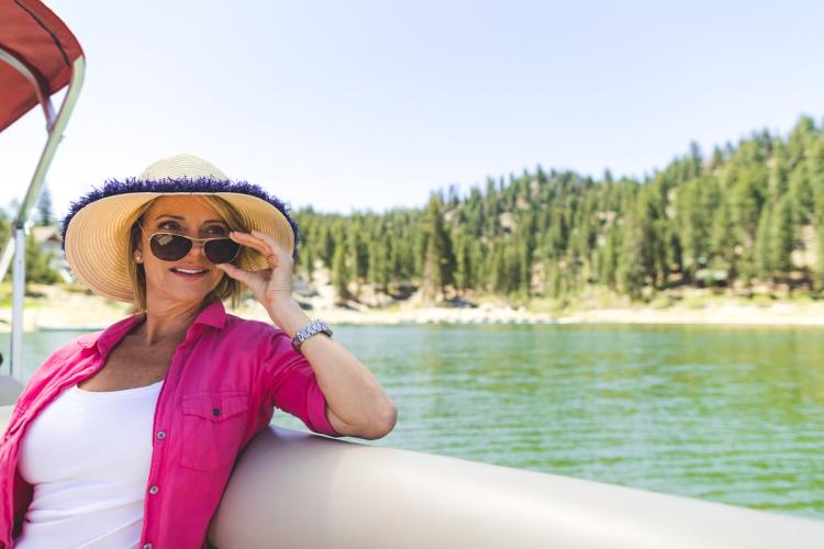 Una mujer en una barca pontón: sostiene sus gafas de sol y mira hacia donde no se ve; detrás de ella se divisan el lago y los árboles.