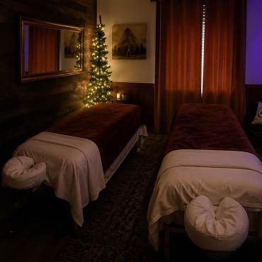 Dos camillas de masaje largas en una sala suavemente iluminada con luz ambiental; las camillas cuentan con fundas mullidas y reposacabezas.