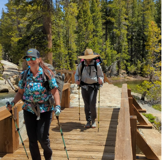 A female and male taking a hiking trail onto a bridge, they have hiking sticks and backpacks .