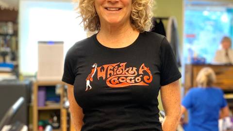 A photo of a Pilates instructor, her hands are behind her back and she has a welcoming smile