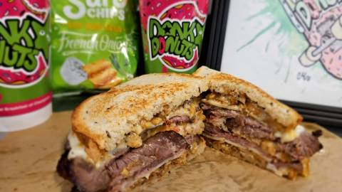 Foto transversal del sándwich caliente «The Lockness Muenster» de Dank Donut; al fondo se ven bebidas calientes.