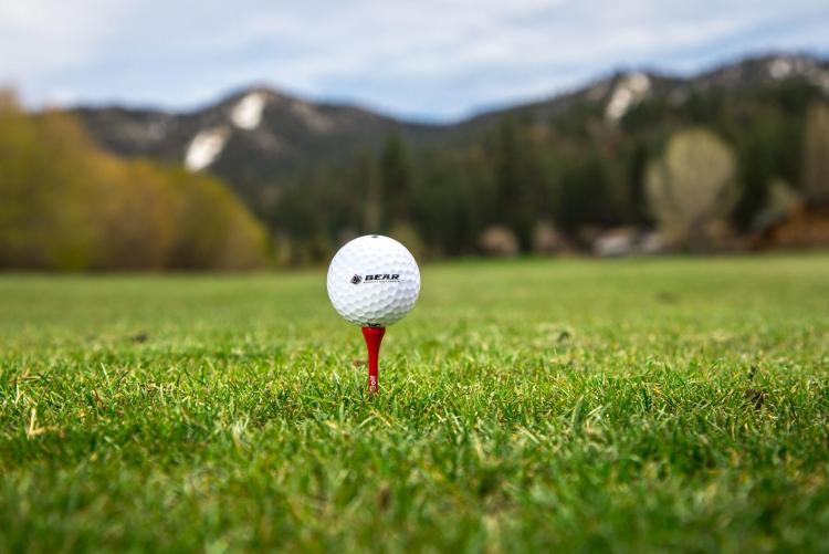 Una pelota de golf blanca con el logotipo de BBMR se posa en un tee rojo sobre hierba verde. En el fondo se ven montañas difuminadas.