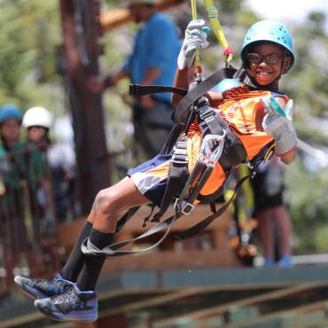 Una foto de un niño atado firmemente para hacer tirolesa con Action Zipline Tours, lleva guantes gruesos y casco.
