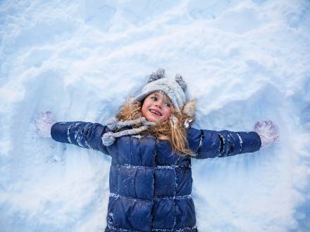 Una foto de una niña con una chaqueta de nieve azul haciendo un ángel en la nieve blanca y esponjosa mientras sonríe feliz.