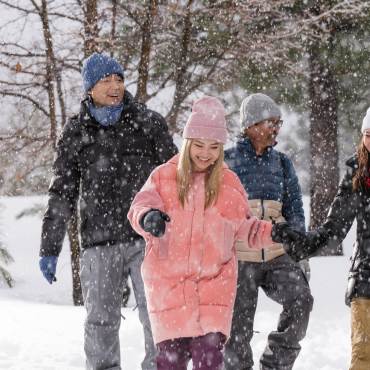 Un grupo de cuatro personas abrigadas en la nieve, con copos cayendo a su alrededor y árboles verdes detrás de ellos.