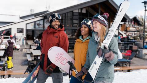 Tres amigos con ropa de invierno, sonriendo con esquís y tablas de snowboard en una estación de esquí nevada.