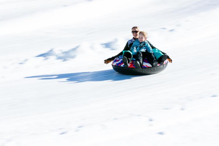 Una foto de una madre y su hijo bajando a toda velocidad por una colina cubierta de nieve montados en un flotador, bien abrigados y sonrientes.