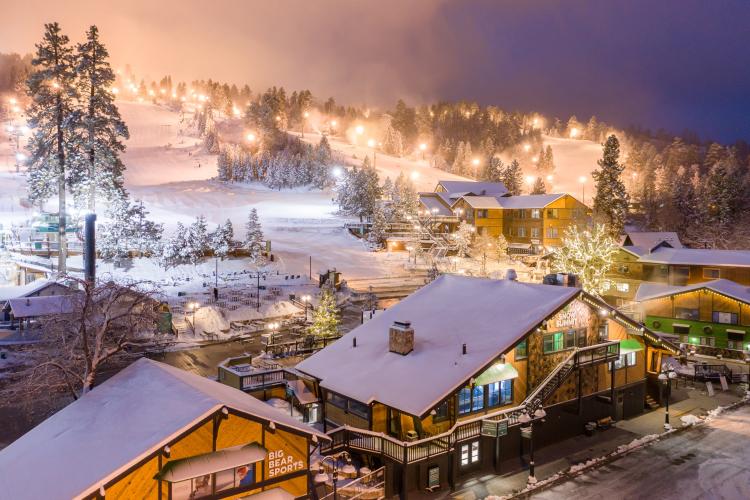 Snow Summit se ilumina por la noche para las sesiones de esquí nocturno durante la temporada de invierno. La nieve cubre los edificios y los árboles.