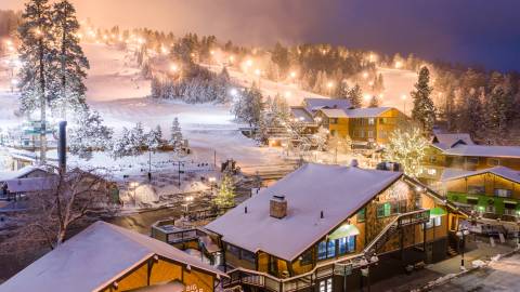 Snow Summit se ilumina por la noche para las sesiones de esquí nocturno durante la temporada de invierno. La nieve cubre los edificios y los árboles.