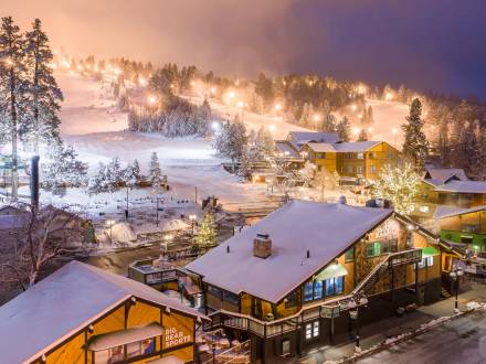 Snow Summit se ilumina por la noche para las sesiones de esquí nocturno durante la temporada de invierno. La nieve cubre los edificios y los árboles.