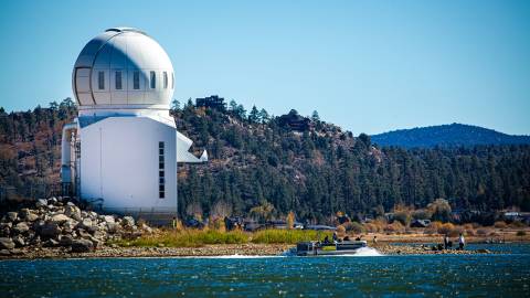 La cúpula blanca del observatorio se alza sobre la orilla del lago Big Bear. Un barco pontón navega cerca.