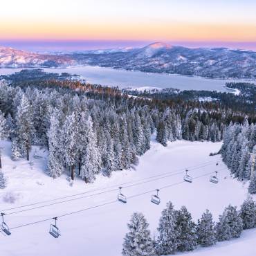 Una foto aérea de las colinas de esquí cubiertas de nieve, el lago Big Bear y las montañas reflejando suavemente la puesta de sol.