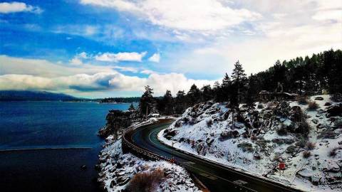 Conducción invernal en Big Bear Lake California con una carretera llena de nieve y viento entre cielos azules y árboles de hoja perenne
