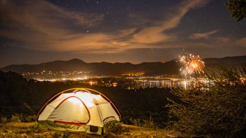 Una foto de una tienda de campaña en una colina con una luz que la ilumina por la noche, con vistas a los fuegos artificiales en Big Bear Lake, California.