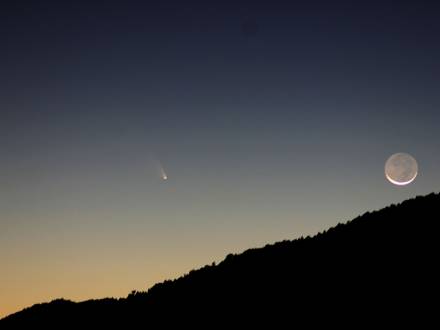 Foto de una montaña en silueta, la luna está en cuarto menguante mientras un cometa pasa a lo lejos.