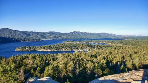 Vista desde lo alto de un acantilado de un bosque, un lago y una cadena montañosa en Big Bear, California, en un día soleado.