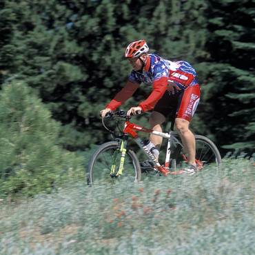 Un ciclista de montaña con una equipación de color rojo brillante recorre un sendero para bicicletas en Big Bear Lake, California. El frondoso y verde bosque le rodea.