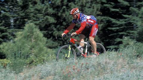 Un ciclista de montaña con una equipación de color rojo brillante recorre un sendero para bicicletas en Big Bear Lake, California. El frondoso y verde bosque le rodea.