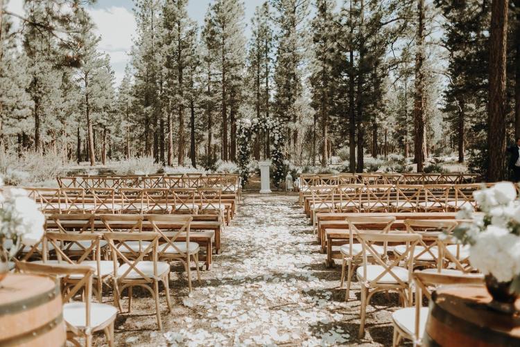 Sillas de madera en hileras en una boda en el bosque de Big Bear. Flores blancas sobre barriles de vino en primer plano.