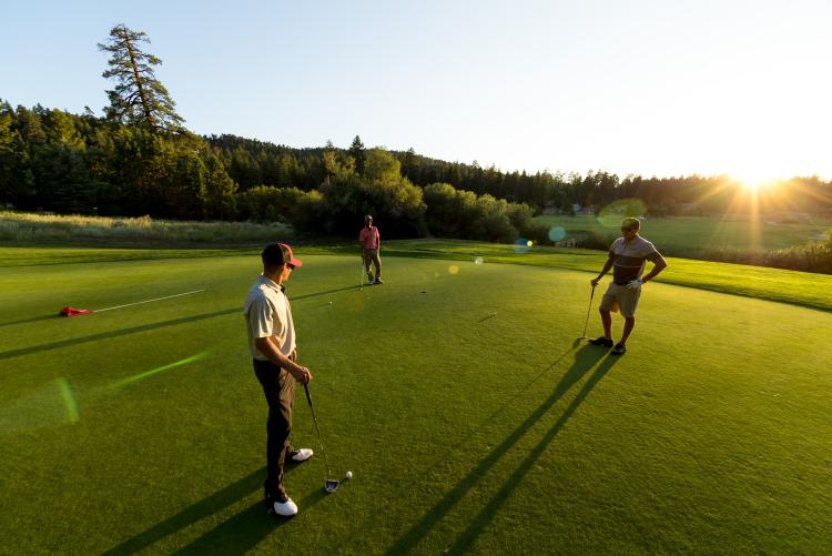 Un grupo de tres hombres jugando al golf en el campo de Bear Mountain, en un terreno llano y verde; se ven los árboles y el sol resplandeciente.