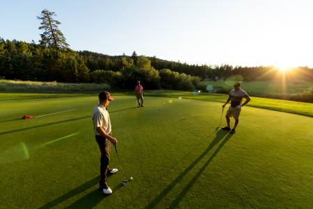 Un grupo de tres hombres jugando al golf en el campo de Bear Mountain, en un terreno llano y verde; se ven los árboles y el sol resplandeciente.