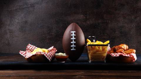 Un balón de fútbol sentado en una mesa de madera con aperitivos como patatas fritas y alitas de pollo. Imagen de archivo.
