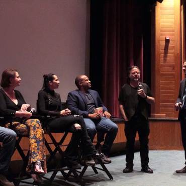 Un grupo de directores de cine sentados en sillas, escuchando a un presentador que habla por un micrófono durante un festival de cine.