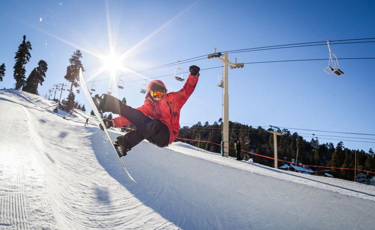 Una foto de un esquiador de snowboard en pleno salto desde una pista de half-pipe: el sol brilla detrás de la persona y sobre la pista de snowboard.