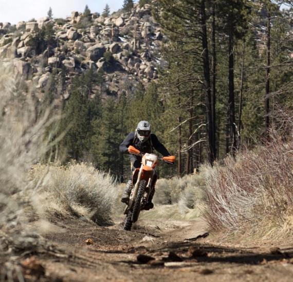 Un motociclista recorre un camino de tierra en su moto naranja, detrás de él hay montañas rocosas y bosques.