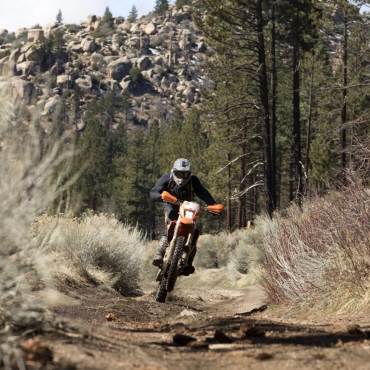 Un motociclista recorre un camino de tierra en su moto naranja, detrás de él hay montañas rocosas y bosques.