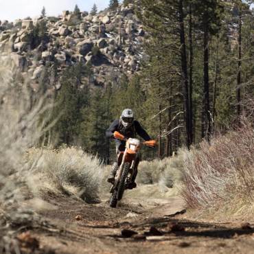 Un motociclista recorre un camino de tierra en su moto naranja, detrás de él hay montañas rocosas y bosques.