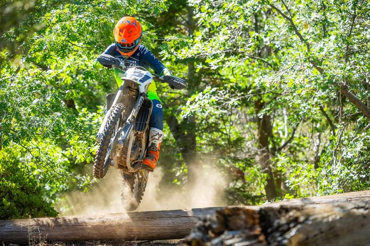 Un piloto de moto de cross captado en pleno vuelo sobre su moto mientras realiza un salto por encima de un tronco en un bosque; detrás de él se ve una nube de polvo.