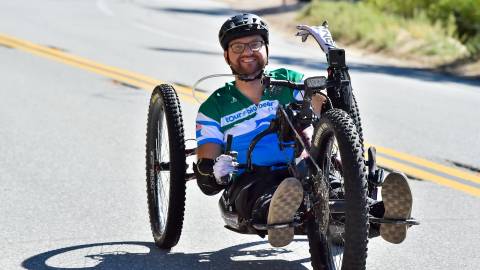 Un hombre conduce un triciclo reclinado sobre el pavimento durante el Tour de Big Bear. Saluda a la cámara y sonríe.