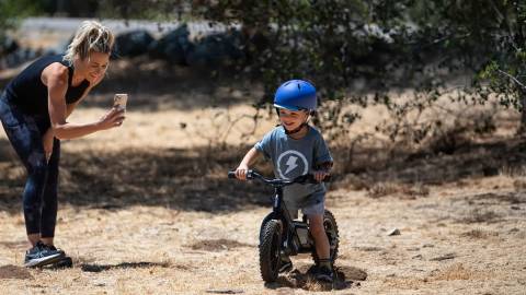 Un niño pequeño que va en una bicicleta Stacyc y lleva un casco azul; su madre está de pie a un lado grabándole.
