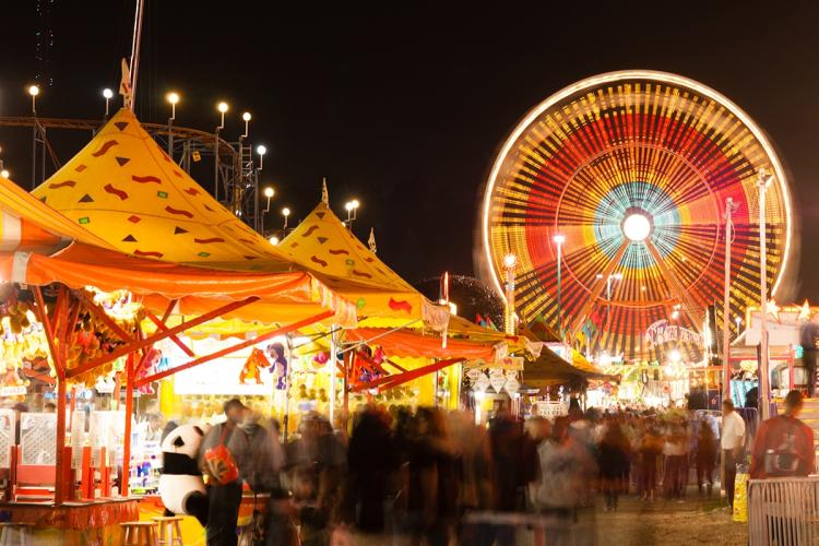 Una gran feria con una noria y puestos de vendedores; la noria está iluminada con colores vivos: rojo, azul y amarillo.