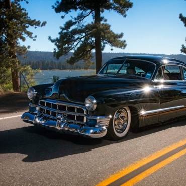 Un coche Hudson antiguo circulando por la carretera de Big Bear Lake; al fondo se divisan las montañas, los árboles y el lago.