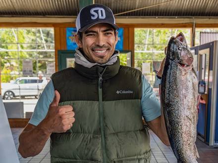 Un hombre con una chaqueta de pesca que sostiene una trucha grande mientras levanta el pulgar y sonríe durante el evento «Fishin' For $50K».