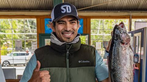Un hombre con una chaqueta de pesca que sostiene una trucha grande mientras levanta el pulgar y sonríe durante el evento «Fishin' For $50K».