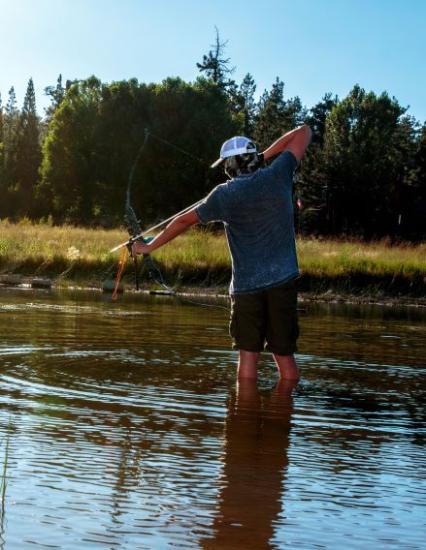 A male with a blue shirt and white hat is wading in water while aiming his bow at the water during the Countin' Carp event.
