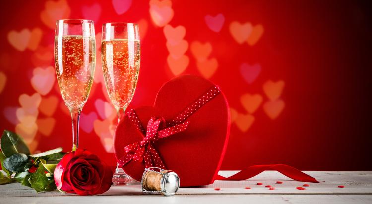 Dos copas de champán sobre una mesa con una caja de bombones en forma de corazón y una rosa roja en una imagen temática de San Valentín.