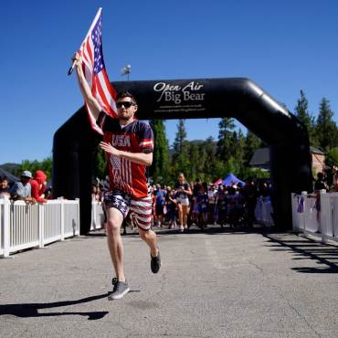 Un corredor porta una bandera estadounidense durante la carrera Big Bear Lake 4th of July Fun Run. Es un día de pájaro azul y los aficionados le animan.