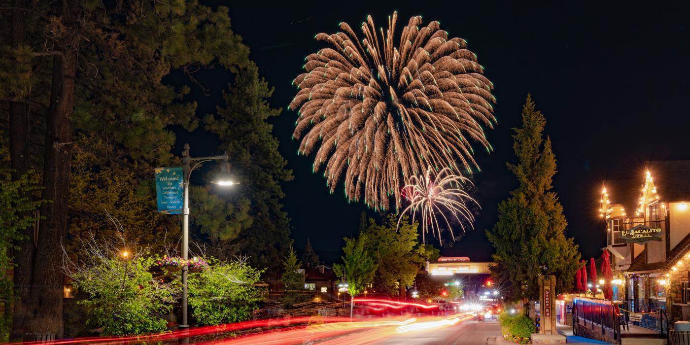 Una larga exposición de luces de cola rojas en The Village en Big Bear Lake. Fuegos artificiales del 4 de julio son visibles en el cielo nocturno.